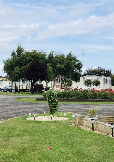 A grass section and holiday homes in the background surrounded by well kept gardens with red flowers